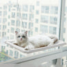 White cat resting on a hanging cat window hammock attached to a glass window with suction cups for birdwatching and sunbathing.